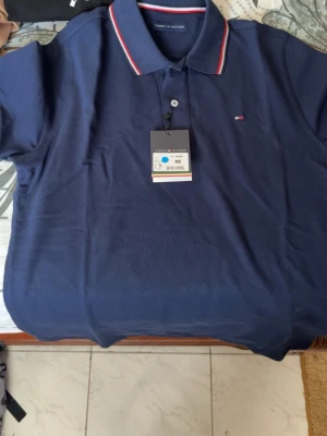 Marinblå pikétröja Tommy Hilfiger - Klassisk marinblå pikétröja från Tommy Hilfiger med krage och knappar framtill. Tröjan har röda och vita ränder på krage och ärmslut samt den ikoniska loggan broderad på bröstet. Perfekt till jeans eller chinos för en clean look.