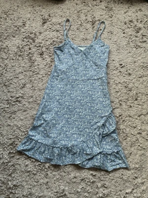 Blå blommig omlottklänning volang - Söt blå kortklänning med små vita blommor, omlottmodell och volangkant nertill. Klänningen har smala axelband och är tillverkad i ett mjukt, lätt material som passar perfekt till varma dagar. Perfekt för dig som gillar en romantisk och avslappnad stil.