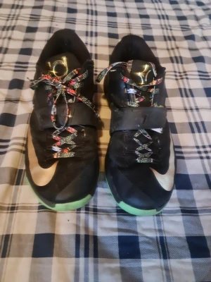 Nike KD 7 sneakers svart/guld - Snygga Nike KD 7 sneakers i svart med guldiga detaljer och en stor Swoosh på sidan. Skorna har färgglada mönstrade skosnören och grön sula. Ovandelen är i mesh och syntetmaterial, med kardborreband över vristen för extra support.