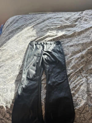 Svarta skinnbyxor med utsvängda ben - Snygga svarta skinnbyxor med utsvängda ben och hög midja. Byxorna har en stilren look och passar perfekt till en trendig outfit. Materialet ger en cool och edgy känsla som lyfter vilken stil som helst.