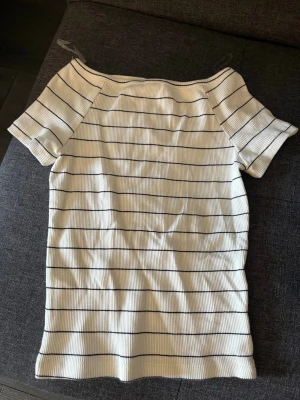 Vit ribbad randig topp - Säljer en vit ribbad topp med svarta horisontella ränder. Toppen har korta ärmar och en rak, något bredare halsringning. Materialet är stretchigt och ger en bekväm passform. Perfekt till jeans eller kjol för en enkel och snygg look. Typ aldrig använt men nu är den lite liten.