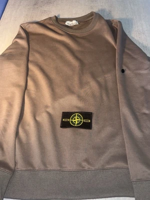 Beige sweatshirt från Stone Island - Snygg beige sweatshirt från Stone Island med klassisk rund halsringning och ribbade muddar. Tröjan har den ikoniska Stone Island-loggan på ärmen i svart och gult. Perfekt för en avslappnad och trendig stil.