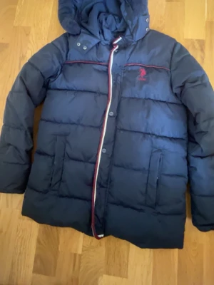 Mörkblå dunjacka från USPA - Snygg mörkblå dunjacka från USPA med röd logga på bröstet och röda detaljer längs dragkedjan och bröstet. Jackan har huva och knäppning med både dragkedja och knappar. Perfekt för kalla vinterdagar.