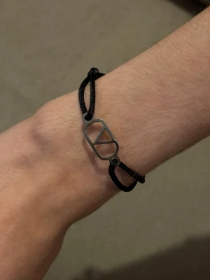 Svart armband med V-logo - Stilrent armband med svart justerbart snöre och en silverfärgad metallbricka med V-logga. Perfekt accessoar för dig som gillar minimalistisk design och vill ha något som sticker ut lite extra. Passar både tjejer och killar.
