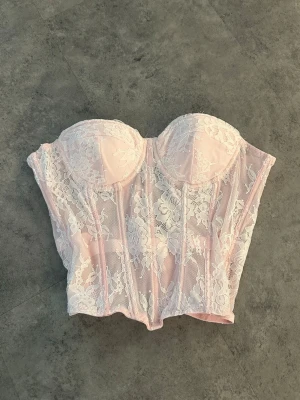 Rosa spetskorsett med snörning - Säljer en superfin rosa korsett med vit spets och vadderade kupor. Den har snygg snörning i ryggen och transparent mesh med blommigt spetsmönster. 