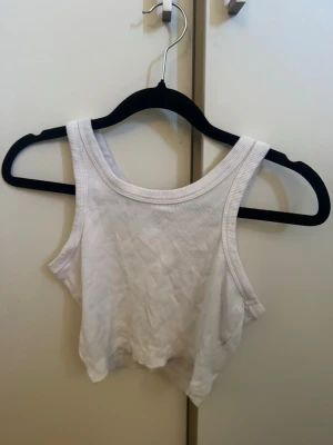 Vit ribbad croppad linnetopp - Säljer en enkel och stilren vit croppad linnetopp med ribbad struktur. Toppen är ärmlös och har breda axelband samt rund halsringning. Perfekt att matcha med höga jeans eller kjol för en avslappnad look.