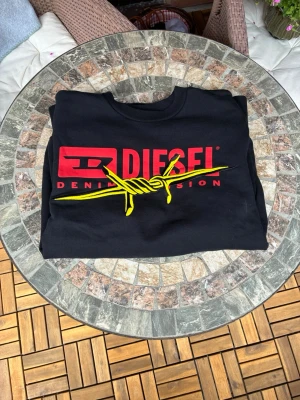 Svart sweatshirt från Diesel XS/S - Cool svart sweatshirt från Diesel med stort rött och gult tryck framtill. Loggan har en taggtrådsgrafik och texten 'DENIM DIVISION'. Tröjan har rund hals och ribbade muddar vid ärmslut och nederkant. Perfekt för en streetwear-look.