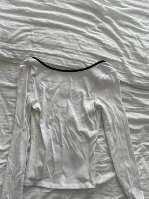 Vit långärmad topp med svart kant - Säljer en vit långärmad topp med en bred, rundad urringning och svart kant runt halsen. Toppen är ribbad och har en slim passform som sitter snyggt på kroppen. 
