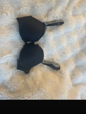 Svart push-up bh från Victoria's Secret - Snygg svart push-up bh från Victoria's Secret med vadderade kupor och spetsdetaljer. Axelbanden har logotyptext och är justerbara för bästa passform. Perfekt för dig som vill ha både komfort och stil.