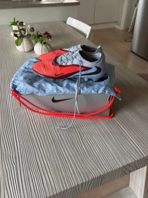 Nike röda och grå fotbollsskor - Säljer ett par Nike fotbollsskor i rött och grått med svarta detaljer. Skorna har snörning och en modern, strömlinjeformad design med dobbar för bra grepp på planen. Tillverkade i syntetmaterial och levereras med tillhörande påse och originalkartong. Det är elites och har med påse