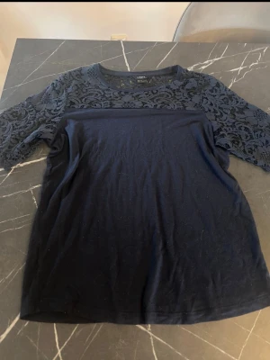Marinblå topp med spetsdetaljer - Snygg marinblå topp från Lindex med spets över axlar och ärmar. Toppen har rund halsringning och halvtransparent spetsmönster upptill samt korta ärmar. Perfekt för dig som gillar feminina detaljer och enkel stil.