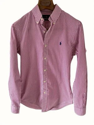 Rödvit randig skjorta från Ralph Lauren - Klassisk skjorta från Ralph Lauren i rödvitt randigt mönster. Skjortan har lång ärm, knappar framtill och en broderad logga på bröstet. Tillverkad i bomull och har en tidlös, normal passform. Perfekt för en clean och stilren look.