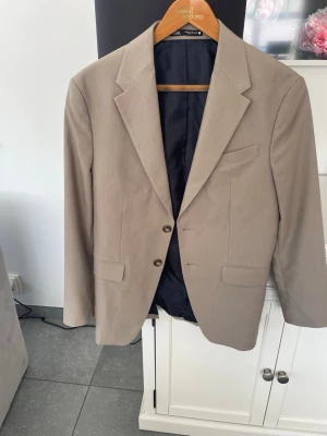 Beige kavaj från Zara herr - Stilren beige kavaj från Zara herr med två knappar framtill, klassisk krage och tre fickor. Kavajen har en slimmad passform och är helfodrad med mörkt innerfoder. Perfekt för en dressad look.