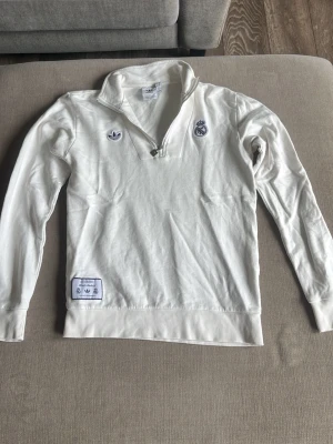Vit halvzip tröja Adidas Real Madrid - Säljer en vit långärmad halvzip tröja från Adidas med Real Madrid-logga på bröstet. Tröjan har ståkrage, broderade detaljer och en patch nertill. Perfekt för dig som gillar sportig stil och vill representera ditt favoritlag.