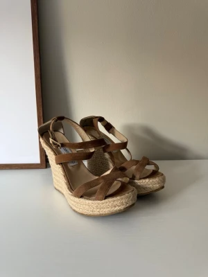 Bruna kilklackade sandaler Jimmy Choo - Snygga bruna sandaler från Jimmy Choo med kilklack i flätad jute och remmar i mockaimitation. Justerbart spänne vid ankeln och öppen tå. Perfekta för sommaren och ger extra höjd med sin stabila platå. Klackhöjd: 12cm 
