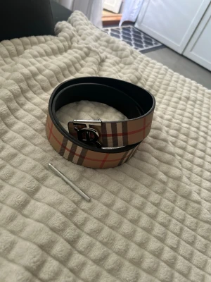 Burberry bälte med klassiskt rutmönster - Snyggt Burberry bälte i skinn med det ikoniska beigea rutmönstret och silverfärgat spänne med logga. Perfekt accessoar för att lyfta din outfit med en touch av lyx och stil.