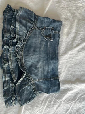 Jeanskjol med volangdetaljer - Säljer en blå jeanskjol med volangkant och dekorativ ficka fram. Kjolen har en kort modell och är sydd i denim med snygga sömmar och tvättad look. Perfekt för dig som gillar en avslappnad men trendig stil.