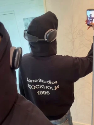 Svart hoodie Acne Studios Stockholm - Svart hoodie från Acne Studios med stor vit text på ryggen där det står 'Acne Studios STOCKHOLM 1996'. Klassisk passform med huva och ribbade muddar. Perfekt för dig som gillar streetwear och vill ha en stilren look.