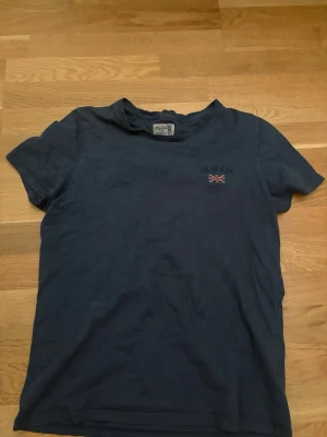 Blå t-shirt från Morris - Snygg mörkblå t-shirt från Morris med liten brittisk flagga och logga på bröstet. Klassisk passform och rund halsringning, perfekt till jeans eller shorts. Tillverkad i mjuk bomull för en skön känsla hela dagen.