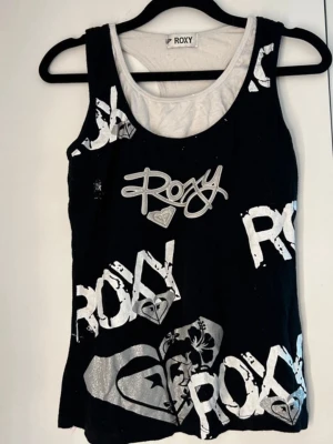 Svart och vit linnetopp från Roxy - Cool svart linnetopp från Roxy med stora vita logotryck och grafiska detaljer framtill. Toppen är ärmlös och har en rund halsringning med kontrasterande vit insida. 