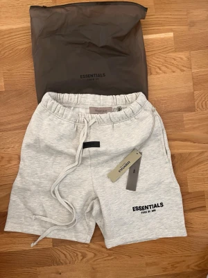 Grå shorts från Essentials Fear of God - Snygga ljusgrå shorts från Essentials Fear of God med elastisk midja och snörning. Mjuk bomullskänsla och logga tryckt på benet. Perfekta för en chill och avslappnad stil.
