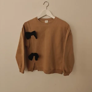 Zara sweatshirt - Beige sweatshirt från Zara med två stora svarta rosetter framtill. Tröjan har rund hals, långa ärmar med lätt puff och ribbade muddar. Mjuk insida och liten slits nertill. Perfekt för dig som gillar unika detaljer!