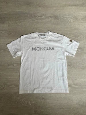 Vit Moncler t-shirt med logga - Snygg vit t-shirt från Moncler med stor logotyp tryckt på bröstet och ett Moncler-märke på ärmen. Klassisk rund hals och korta ärmar. Tillverkad i mjuk bomull för en skön känsla och clean look.