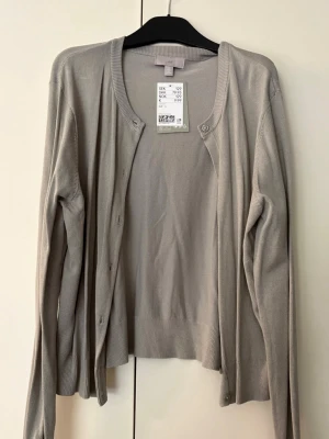 Ljusgrå cardigan från H&M strl.L - Stilren ljusgrå cardigan från H&M med rund halsringning och knäppning framtill. Tillverkad i mjukt, tunt material med ribbade muddar vid ärmslut och nederkant. Perfekt att slänga över en topp för en clean look.
