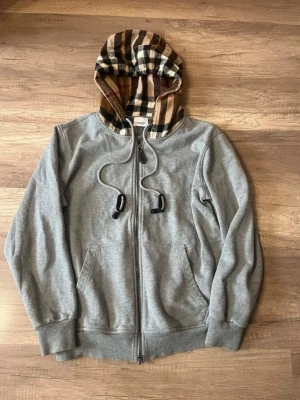Grå hoodie från Burberry med mönstrad huva - Säljer en grå hoodie från Burberry med dragkedja och fickor framtill. Huvan har det klassiska Burberry-rutiga mönstret i beige, svart och rött. Tillverkad i mjuk bomull med lite elastan för extra komfort. Perfekt för en avslappnad och stilren look.