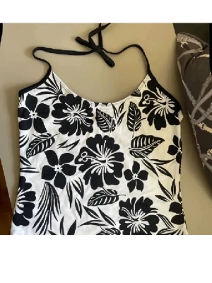 Svartvit blommig linnetopp - Snygg linnetopp med svartvita blommor och tunna axelband som knyts i nacken. Toppen har en croppad passform och är perfekt för varma dagar. Materialet känns lätt och svalt, och mönstret ger en fräsch sommarkänsla.