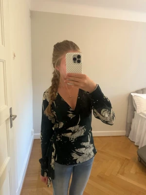 Svart omlottblus med blommönster - Snygg svart omlottblus med stora vita blommor och knytband i midjan. Blusen har djup v-ringning och långa ärmar. Perfekt för dig som gillar en feminin och trendig stil. Materialet känns mjukt och följsamt.