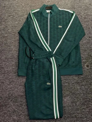 Grön tracksuit äkta - Mörkgrön tracksuit  med vit och grön rand längs ärmar och byxben. Setet har dragkedja på jackan, ståkrage och elastisk midja på byxorna. Diskret Lacoste-logga på både jacka och byxa. Perfekt för chill eller streetwear-vibe.