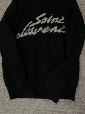 Svart stickad Saint Laurent tröja - Snygg svart stickad tröja från Saint Laurent med vit logotyp i grafisk stil över bröstet. Tröjan har rund halsringning och ribbade muddar vid ärmslut och nederkant. Perfekt för dig som gillar exklusiva och trendiga plagg. Skick 10/10 använt endast fåtal gånger. Storlek S men passar även M
