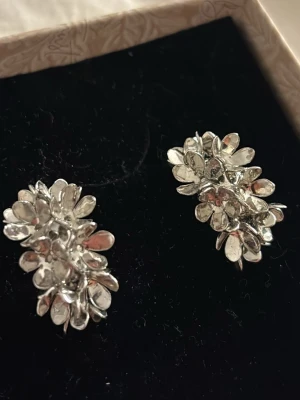 Silvriga blomformade örhängen - Unika örhängen i silverfärg med blomformad design. Varje örhänge består av flera små, glänsande blad som ger ett coolt och iögonfallande intryck. Perfekta för dig som vill ha något annorlunda och snyggt i öronen. 
