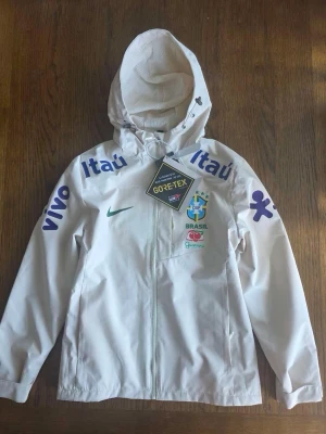 Vit Brasil vindjacka GORE-TEX - Snygg vit vindjacka med Brasil-logga och flera sponsortryck i blått och grönt. Jackan har huva, hel dragkedja och är tillverkad i GORE-TEX-material som står emot vind och regn. Perfekt för dig som gillar sportig stil och vill sticka ut med landslagsvibbar. Det står att det är M storlek men egentligen är den S. Den passar som S och inte M