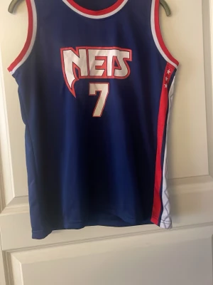Blå Nets basketlinne Durant #7 - Snyggt blått basketlinne från Nets med nummer 7 och Durant på ryggen. Linnet har röda och vita detaljer längs kanterna och sidan, samt tryckt logga framtill. Perfekt för basketfans som vill sticka ut på planen eller på läktaren.