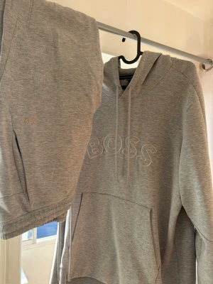Grått loungewear-set från BOSS - Säljer ett grått loungewear-set från BOSS med matchande hoodie och byxor. Hoodien har broderad BOSS-logga på bröstet, dragsko i huvan och stor magficka. Byxorna har resår i midjan och liten BOSS-brodyr. Mjukt och bekvämt material, perfekt för chill dagar.