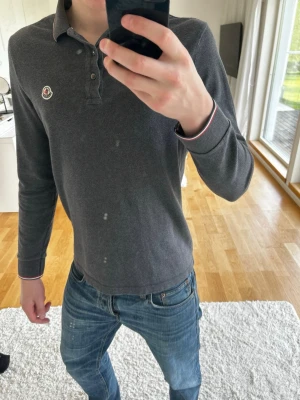 Mörkgrå Moncler långärmad piké - Snygg mörkgrå långärmad pikétröja från Moncler med klassisk krage och knappar framtill. Tröjan har vita och röda detaljer på krage och muddar samt Moncler-logga på bröstet. Perfekt för en clean och stilren look.
