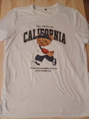 Vit t-shirt med björntryck California - Vit t-shirt med tryck av en söt björn i streetwear och texten 'Los Angeles California'. T-shirten har rund hals och korta ärmar. Perfekt för dig som gillar casual och lekfull stil.