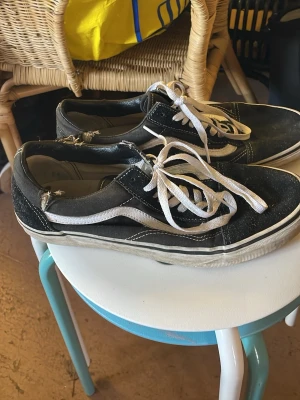 Svarta Vans Old Skool sneakers - Klassiska Vans Old Skool sneakers i svart med vit sula och vit sidostripes. Skorna har vita snören och kontrastsömmar. Ovandelen är i canvas och mocka, vilket ger en skön och avslappnad look.