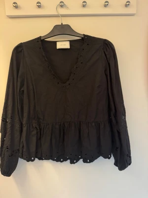 Svart blus med broderade detaljer - Svart blus från neo noir med v-ringning och broderade spetsdetaljer längs halsringning, ärmslut och nederkant. Blusen har lång ärm och en lätt volang nedtill för en feminin look. Perfekt att styla med jeans eller kjol.