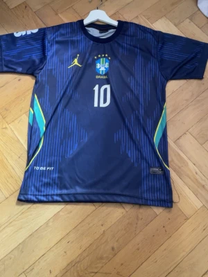 Blå Brasilien fotbollströja Vini Jr - Snygg blå fotbollströja från To Be Fit med Brasilien-emblem och nummer 10 på bröstet. På ryggen står det VINI JR och nummer 10. Tröjan har gula och gröna detaljer på sidorna och är tillverkad i ett lätt, snabbtorkande material. Perfekt för fotbollsträning eller match.
