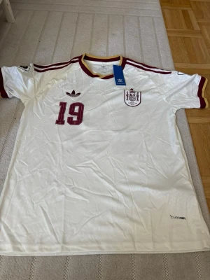Vit Spanien fotbollströja Adidas - Vit Spanien fotbollströja från Adidas med röd-gul krage och röda detaljer. Nummer 19 och Lamine Yamal tryckt på ryggen. Climalite-material som andas, perfekt för träning eller match. Klassisk Adidas-logga och landslagsemblem på bröstet.