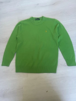 Grön stickad tröja från Polo Ralph Lauren - Snygg grön stickad tröja från Polo Ralph Lauren med rund halsringning och den klassiska lilla gula loggan broderad på bröstet. Tröjan har långa ärmar och ribbade muddar vid ärmslut och nederkant. Perfekt för dig som gillar färg och stilren design.