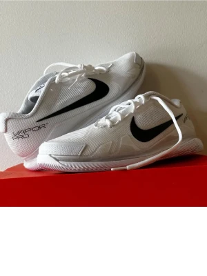 Nike Vapor Pro vita sneakers - Fräscha vita Nike Vapor Pro sneakers med svart swoosh på sidan. Skorna har en låg silhuett och är tillverkade i meshmaterial för bra andningsförmåga. Snygg och sportig design med tydliga detaljer och logga på hälen.