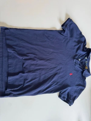 Marinblå piké från Polo Ralph Lauren - Säljer en marinblå pikétröja från Polo Ralph Lauren med röd broderad logga på bröstet. Modellen är Custom Slim Fit och har klassisk krage med knappar. Tillverkad i bomull för en skön känsla och stilren look. Endast använd ett fåtal gånger 