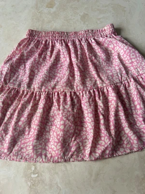 Rosa leopardmönstrad kortkjol - Söt rosa kortkjol med vitt leopardmönster. Kjolen har resår i midjan och volangdetalj som ger en avslappnad och trendig look. Kjolen passar perfekt en varm sommardag.