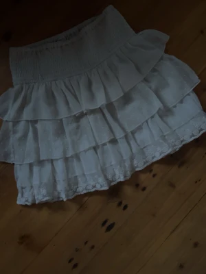 Vit volangkjol med spetsdetaljer - Supersöt vit kortkjol med flera lager volanger och fina spetsdetaljer längst ner. Kjolen har bred resår i midjan för skön passform och är perfekt för sommarens alla outfits. Lätt och luftig känsla.