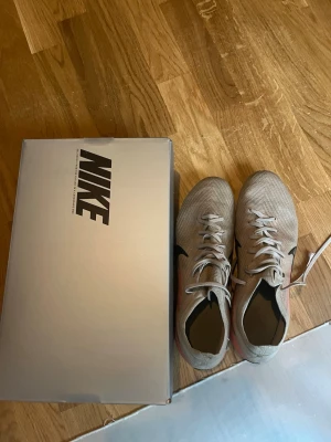 Nike Mercurial Vapor 14 fotbollsskor - Säljer ett par Nike Mercurial Vapor 14 fotbollsskor i beige med svarta och rosa detaljer. Skorna har snörning, låg profil och en lätt ovandel i syntetmaterial för bra bollkontroll. Sulan har dobbar anpassade för spel på gräs. Perfekta för dig som vill ha speed och kontroll på planen.