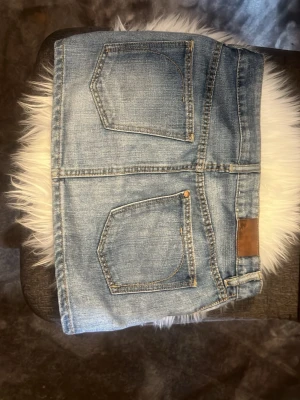 Blå jeanskjol från Juicy Couture - Säljer en klassisk blå jeanskjol från Juicy Couture med fem fickor, bälteshällor och knappstängning fram. Kjolen har en kort modell och är tillverkad i slitstarkt bomullsmaterial med snygga detaljer i guld. Perfekt till alla dina outfits!